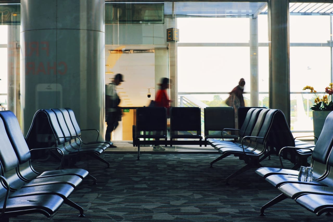 Rows of empty gate seats by tall windows as travelers walk past in an airport