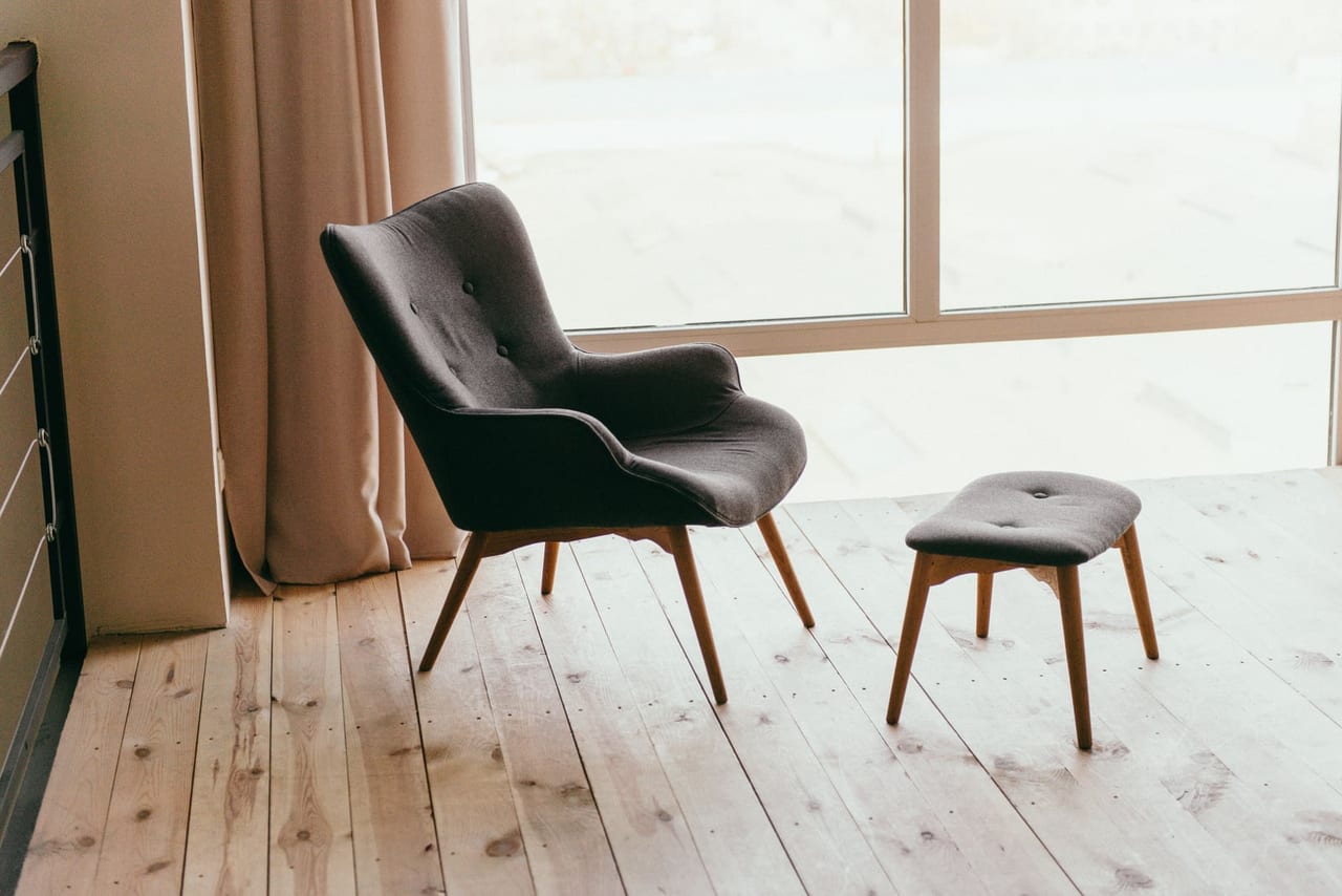 Charcoal armchair and footstool on a wooden plank floor beside a large sunlit window