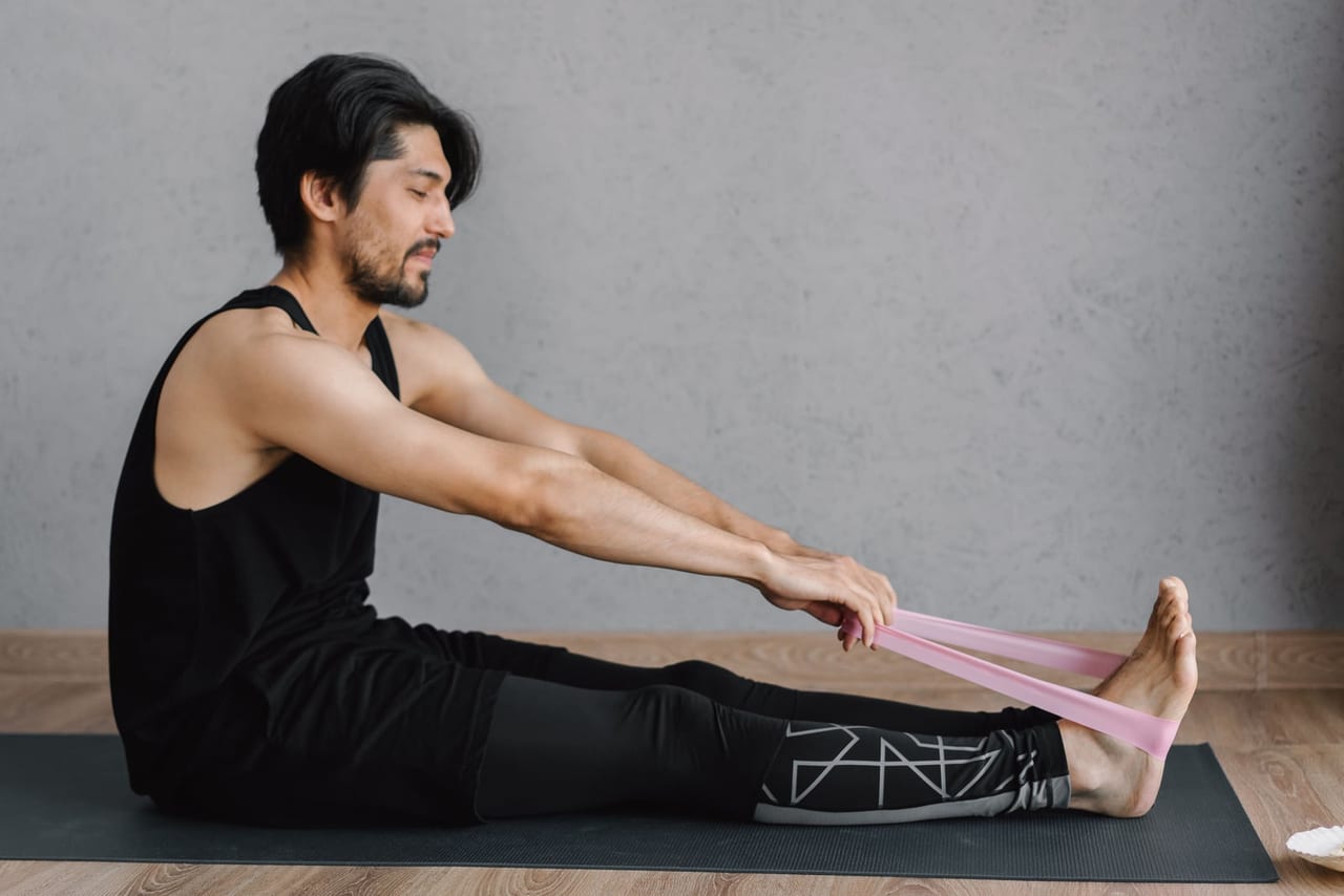 Man seated on a mat holding a pink band looped around his bare feet