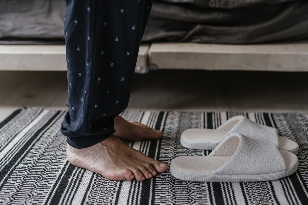Bare feet beside a pair of light gray slippers on a striped woven rug