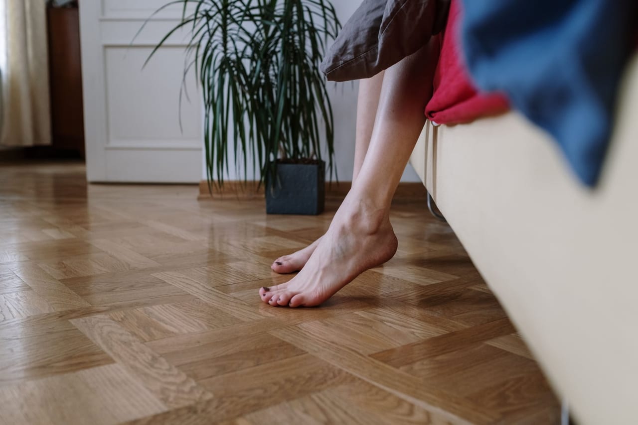Bare feet stepping down from the edge of a bed onto a wooden parquet floor