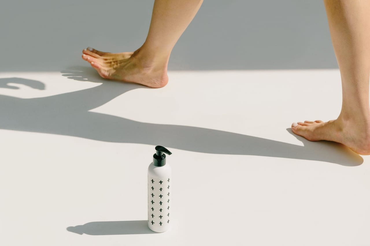 Bare feet stepping across a sunlit white floor beside a white pump bottle