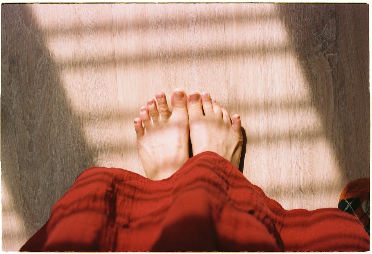 Bare feet resting side by side on a sunlit wooden floor below a red skirt