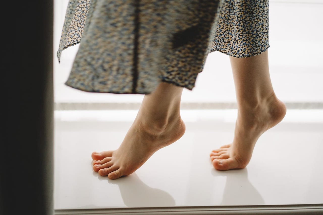 Bare feet rising onto tiptoe on a bright white floor below a long patterned skirt