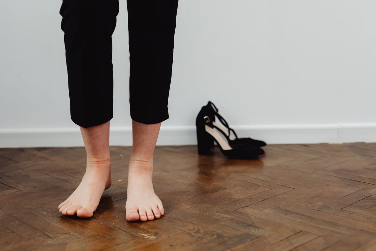Person in black cropped pants standing barefoot on a parquet floor beside black heels