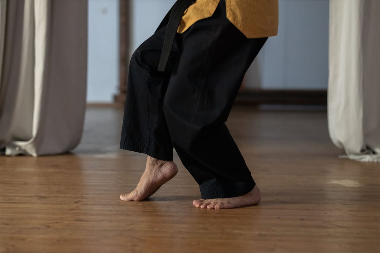 Bare feet on a wooden floor indoors, one heel lifted, under loose black pants