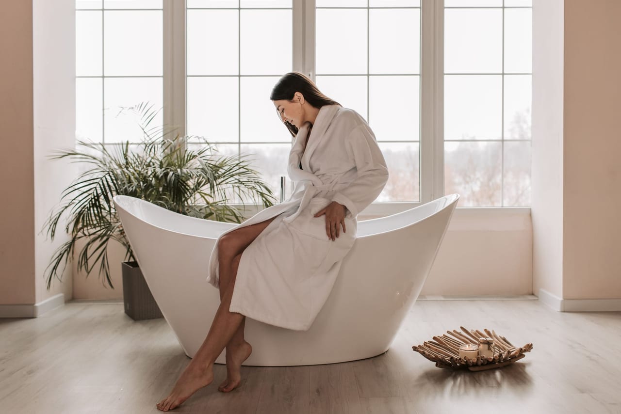 Barefoot woman in a white bathrobe sitting on the rim of a bathtub by tall windows