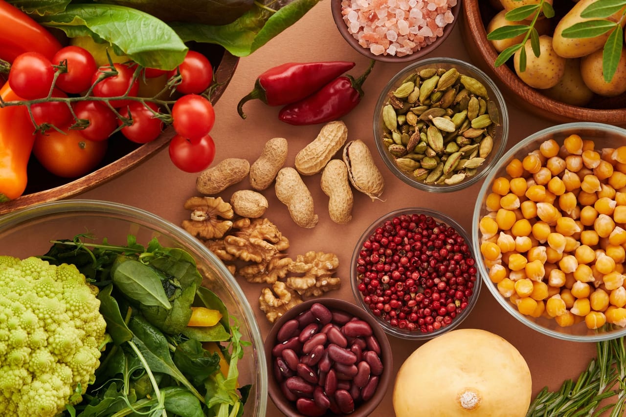 Spinach, kidney beans, chickpeas, walnuts, peanuts and tomatoes in bowls on a tan surface