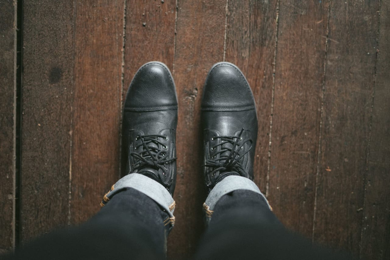 Black leather lace-up boots and cuffed jeans seen from above on a dark wooden floor