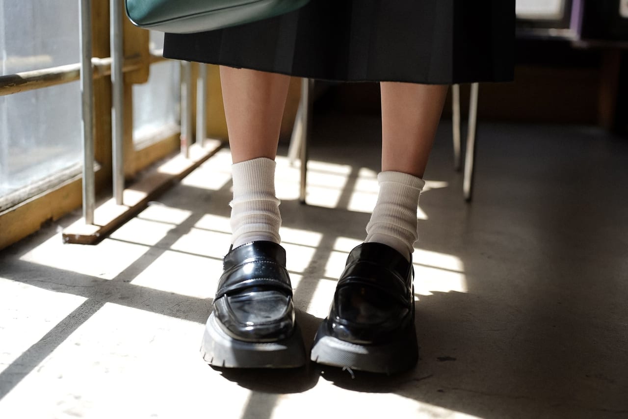 Person standing in black chunky loafers and cream socks on a floor striped with window light
