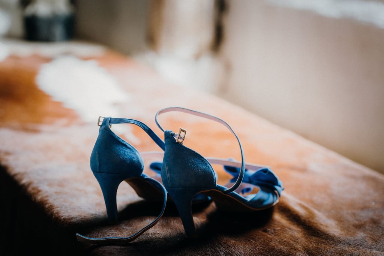 Pair of blue suede ankle-strap heels resting on a cowhide bench in soft light