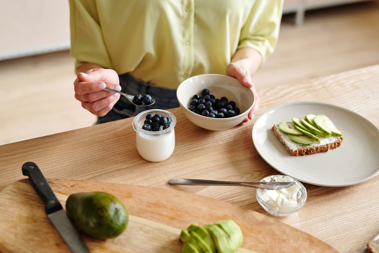 Hands spooning blueberries over a yogurt jar beside a bowl of berries and avocado toast