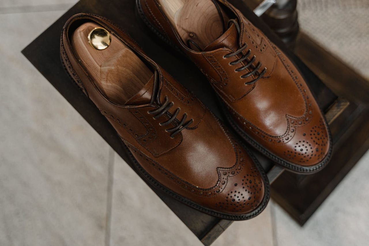 Polished brown leather wingtip brogues with wooden shoe trees inside, on a dark wooden stand