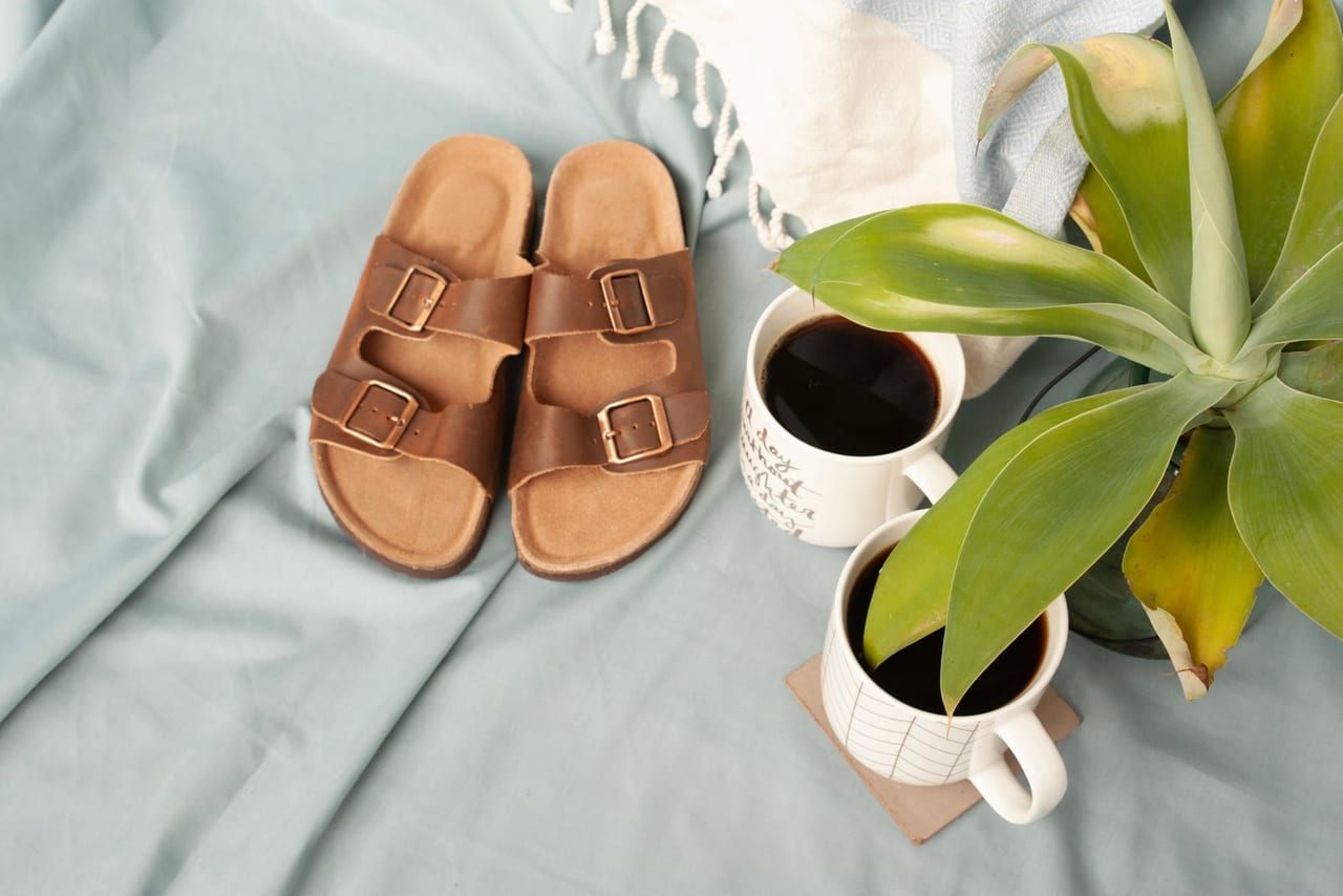 Pair of brown two-strap buckle sandals on a pale blue sheet beside two coffee mugs