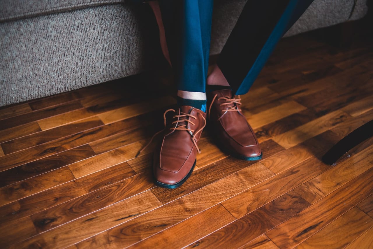 Brown leather lace-up shoes worn with blue trousers on a warm wood floor by a sofa