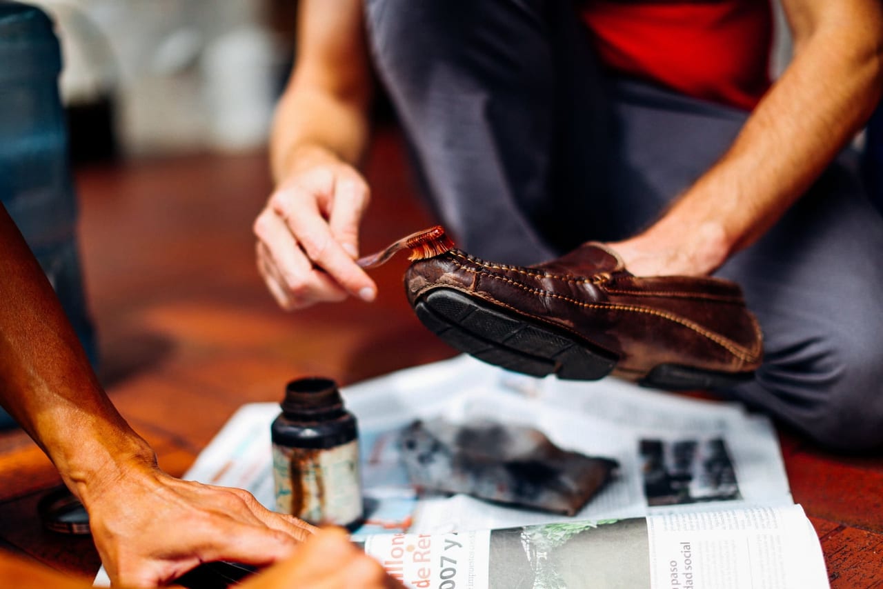 Hands brushing polish onto a brown leather moccasin above newspaper, next to an open jar