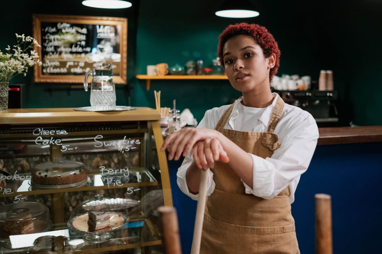 Café worker in a tan apron leaning on a broom handle beside a pastry case