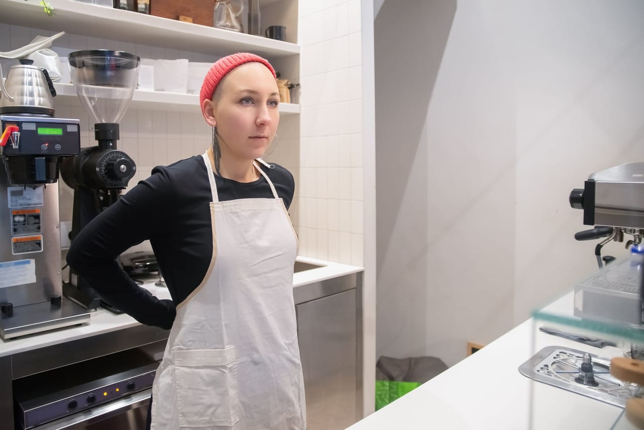 Worker in a white apron and red beanie standing at a cafe counter, hand on hip