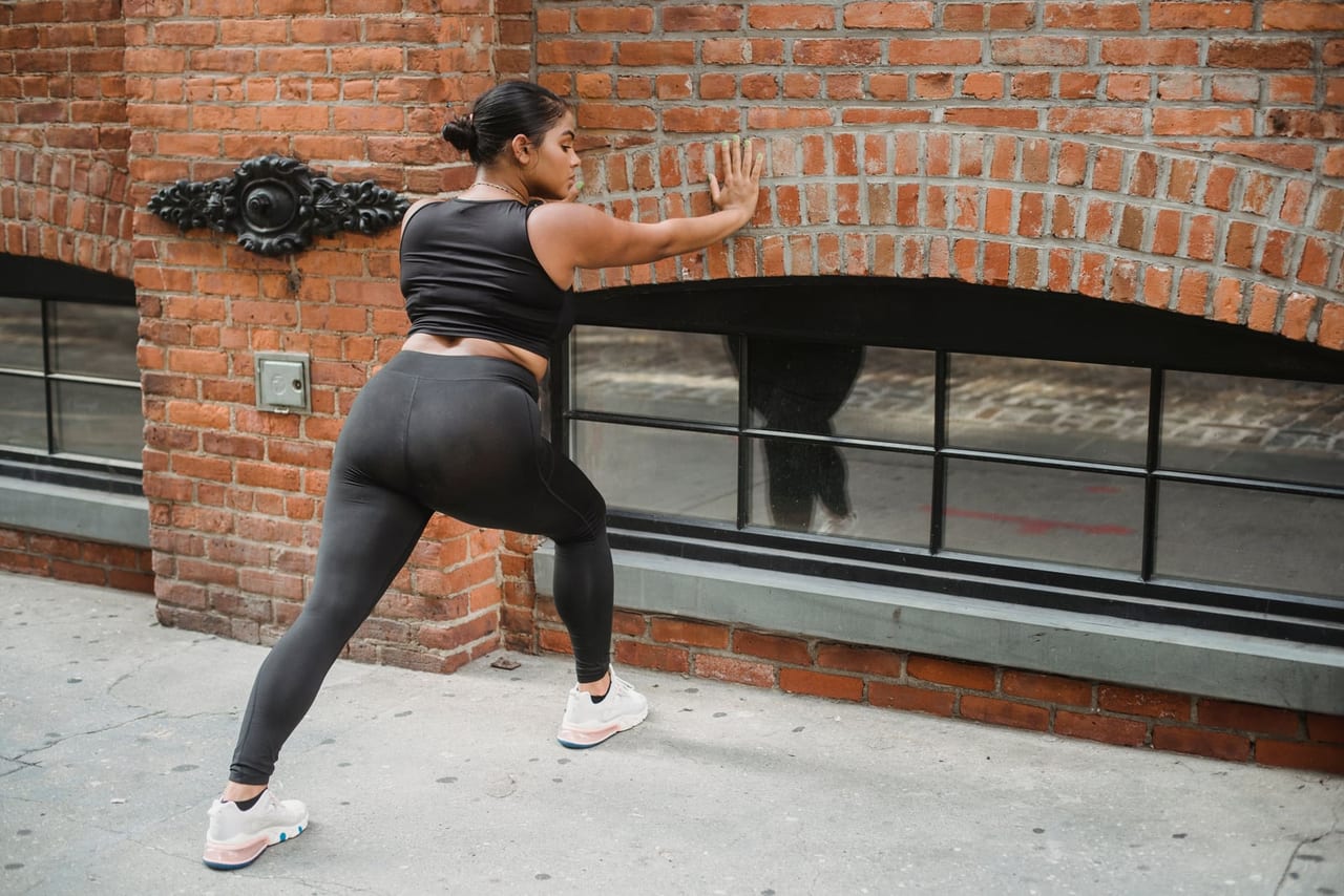 Woman stretching with one hand on a brick wall and one leg stepped back