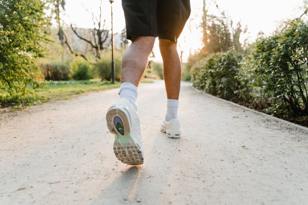 A person's calves and white sneakers seen from behind, mid-stride on a sunlit park path