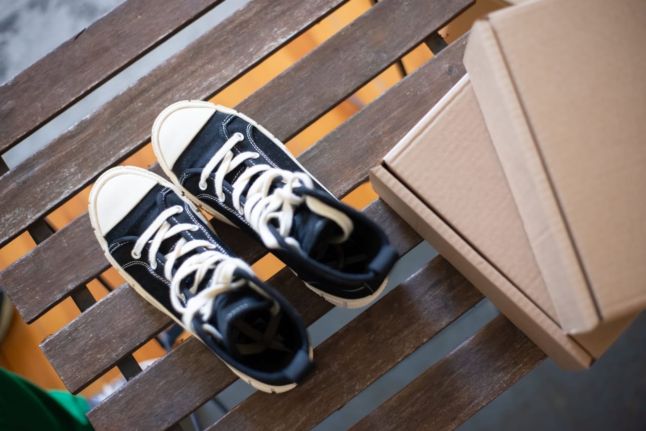 Pair of black canvas high-top sneakers on a slatted wooden bench beside cardboard boxes
