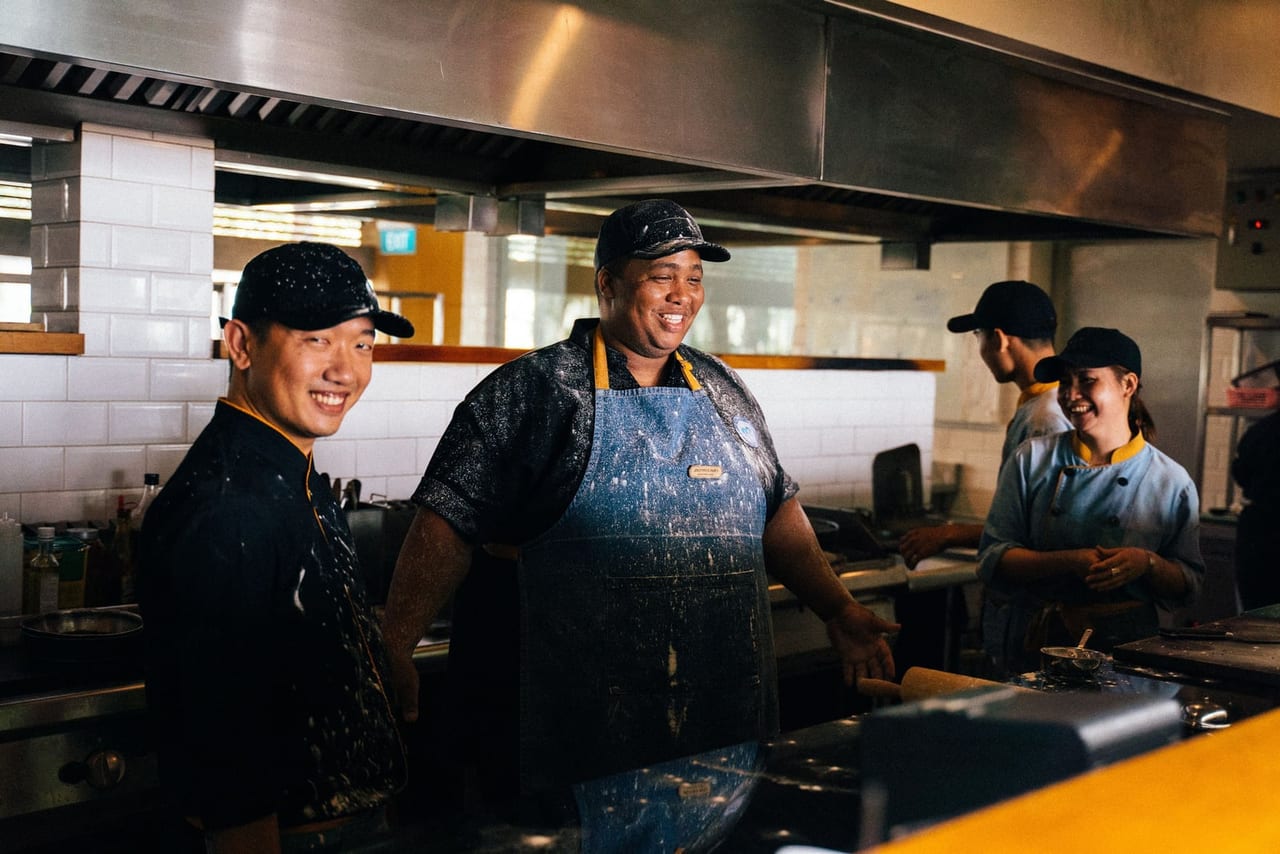 Cooks in caps and aprons laughing together while working in a busy restaurant kitchen