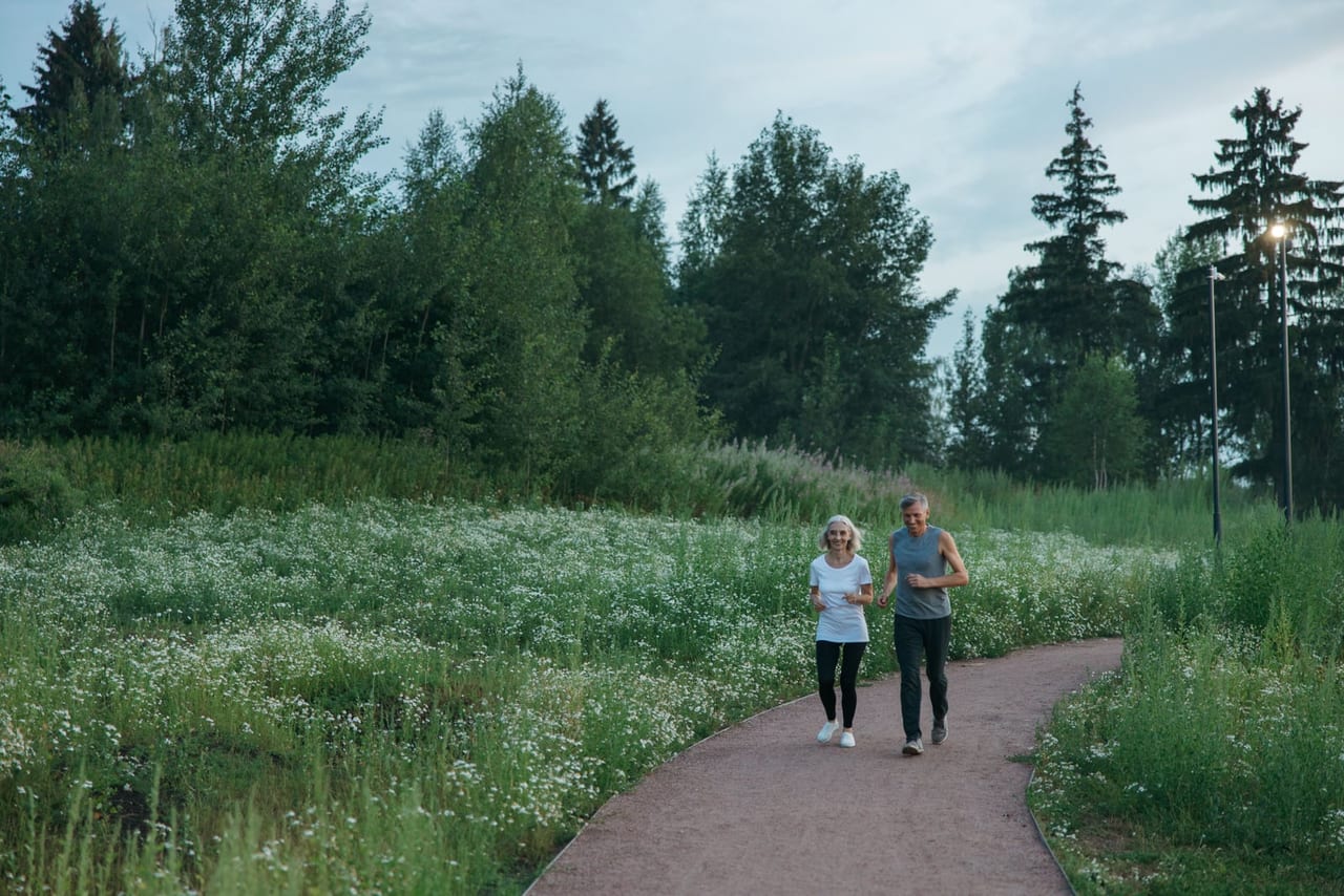 Two older adults jogging side by side along a curving path through a meadow of white wildflowers