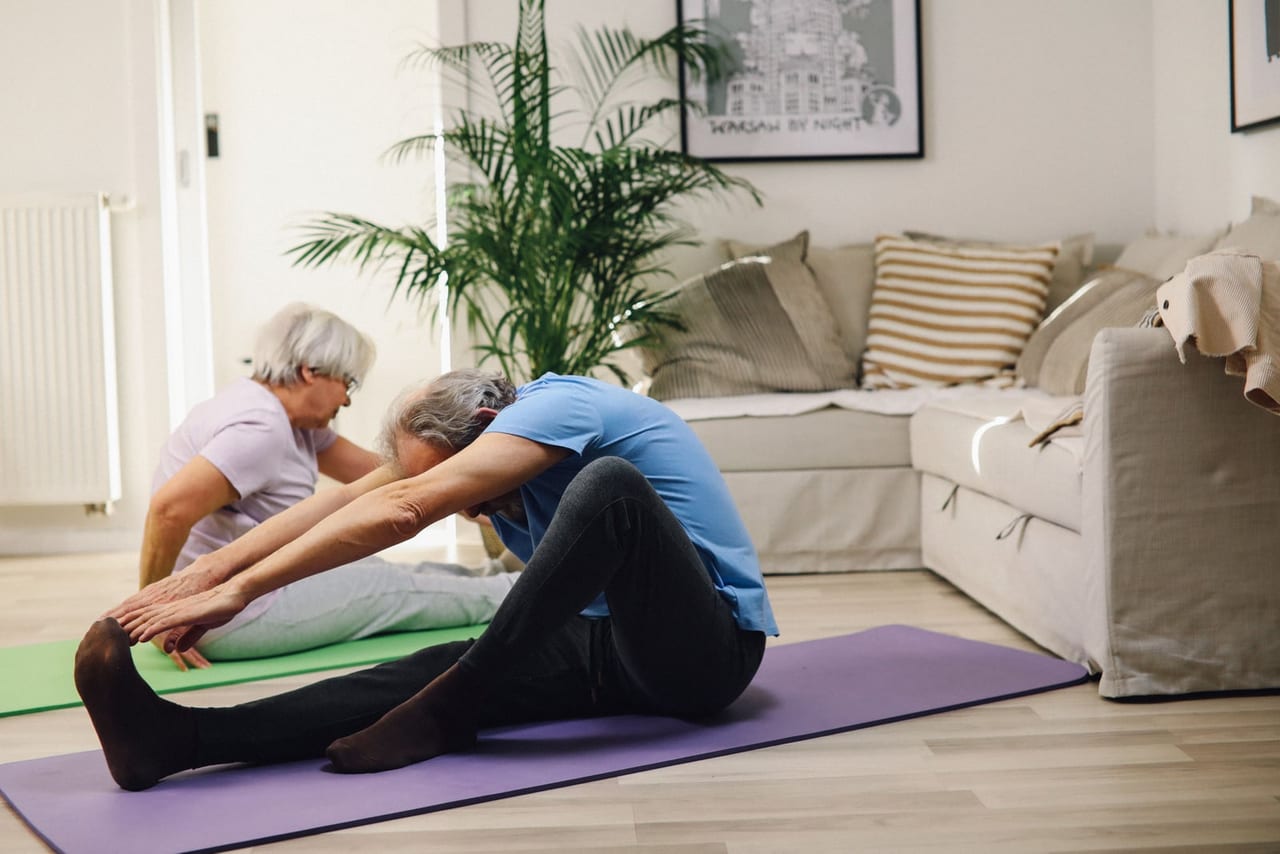 Older couple sitting on yoga mats in a living room, reaching forward toward their feet
