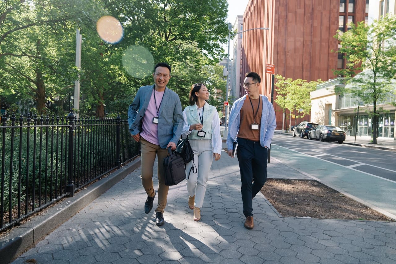 Three people with ID badges and bags walking along a tree-lined city sidewalk in sunlight