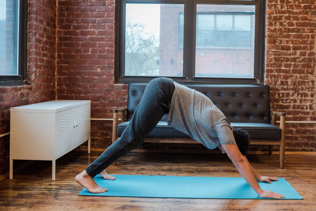 Barefoot man in downward-facing dog on a blue mat in a brick-walled room, heels lifted