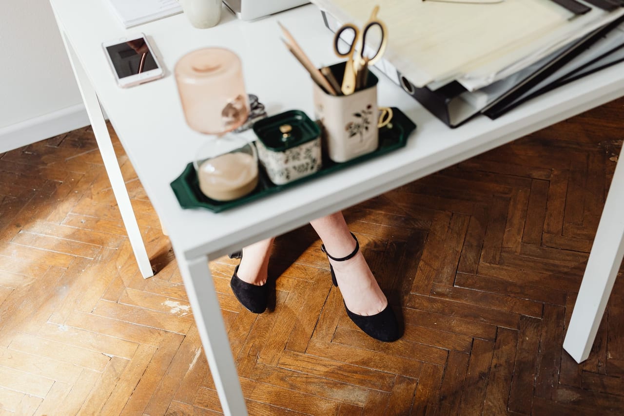 Feet in black strap shoes resting under a white desk on a parquet floor