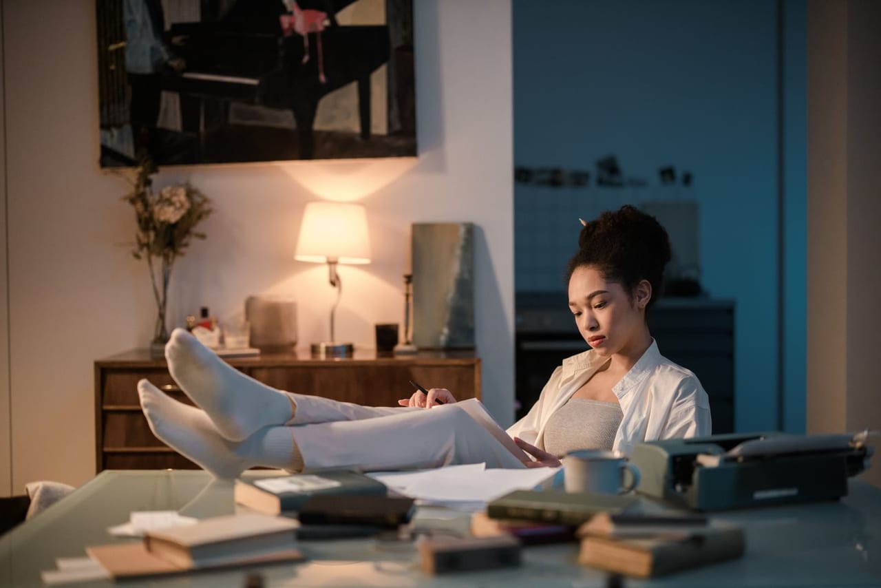 Woman in white socks resting her feet on a desk beside books in lamplight