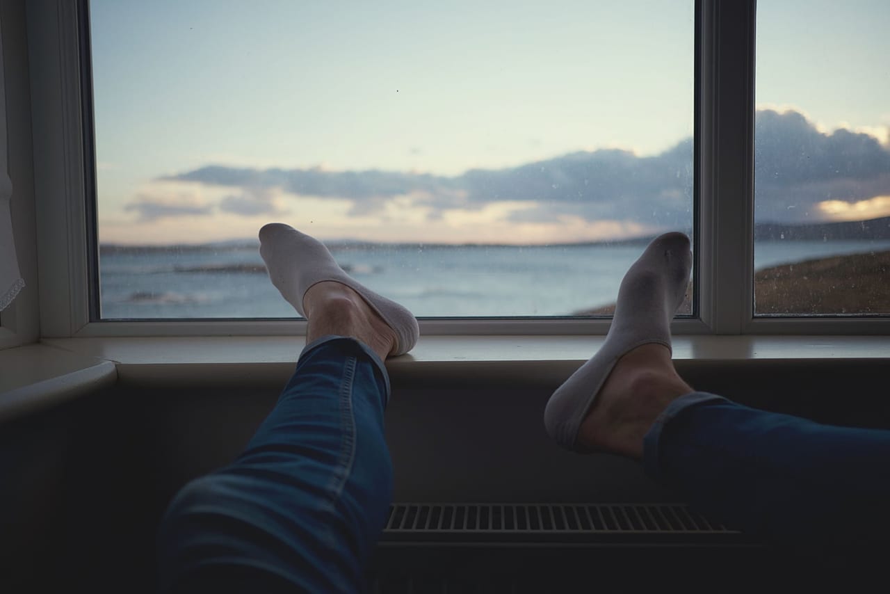 Jeans-clad legs with feet in light socks propped on a windowsill overlooking the sea