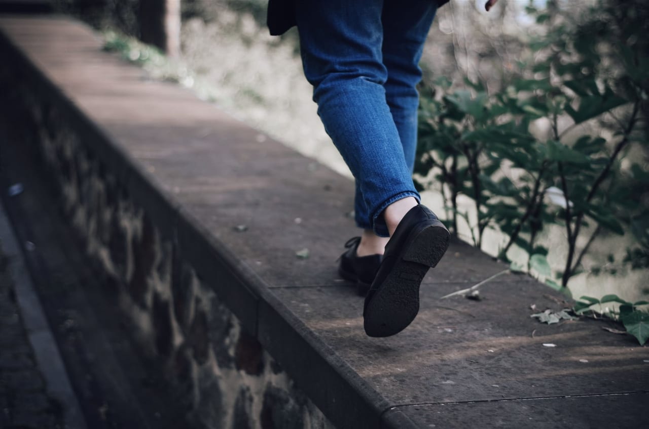 Person in jeans and flat black lace-up shoes stepping along a stone ledge beside plants