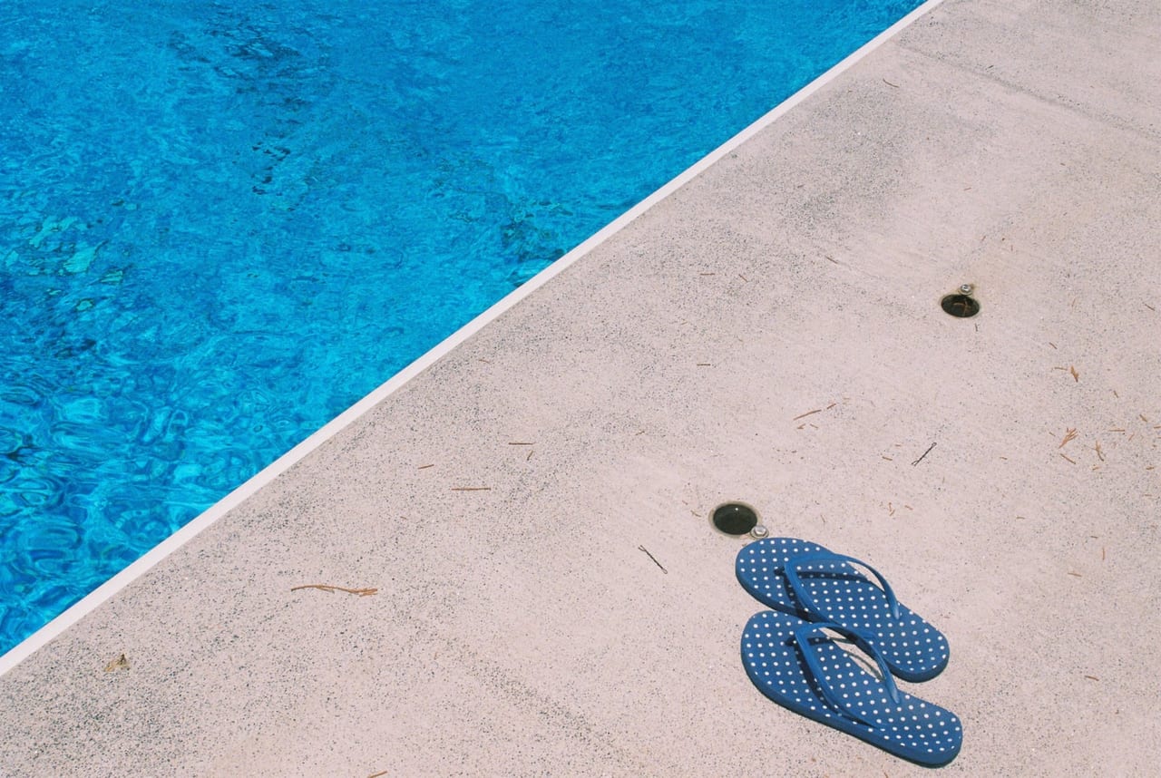 Pair of blue polka-dot flip-flops on a sunlit pool deck beside clear blue water