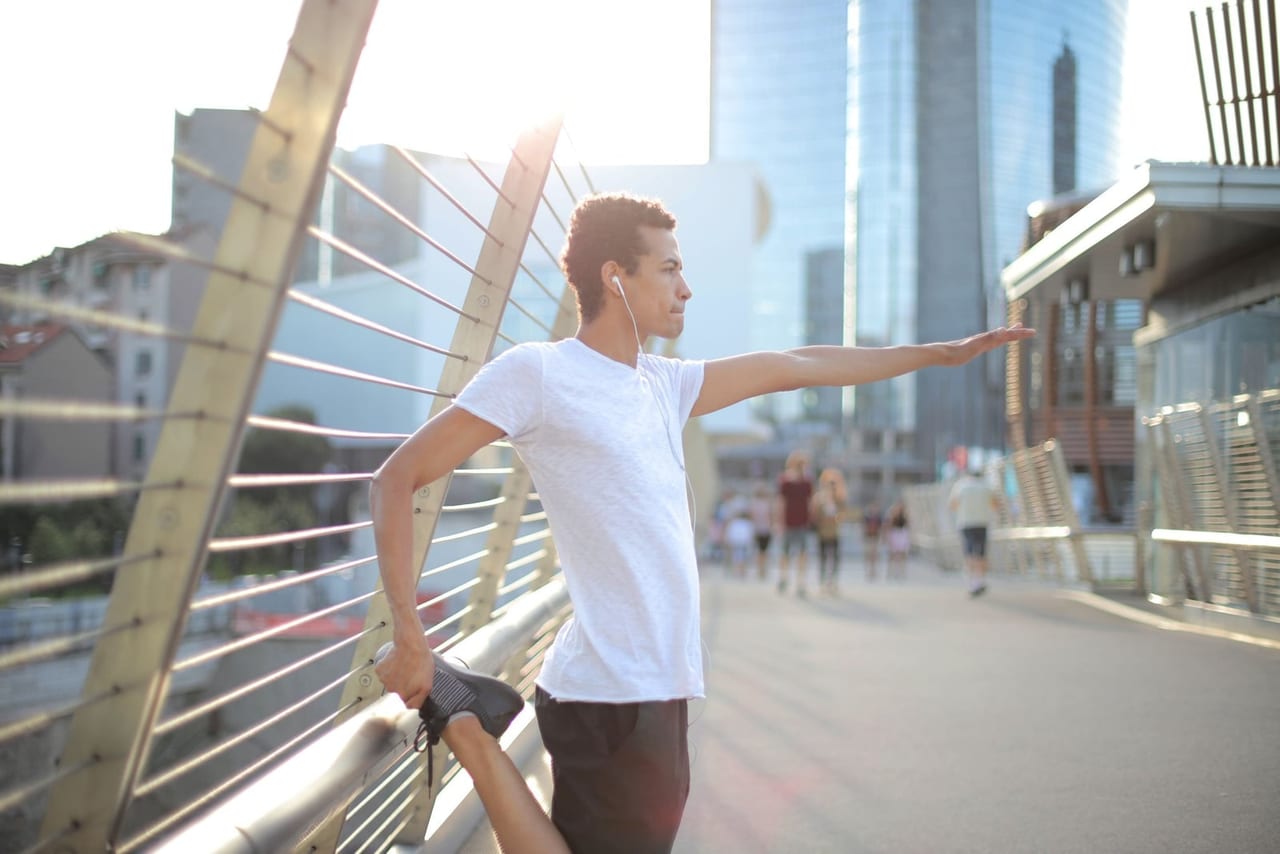 Man in a white T-shirt holding one foot behind him on a sunny city footbridge
