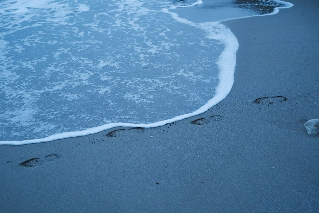 Footprints in wet blue-gray beach sand beside the foamy edge of a wave