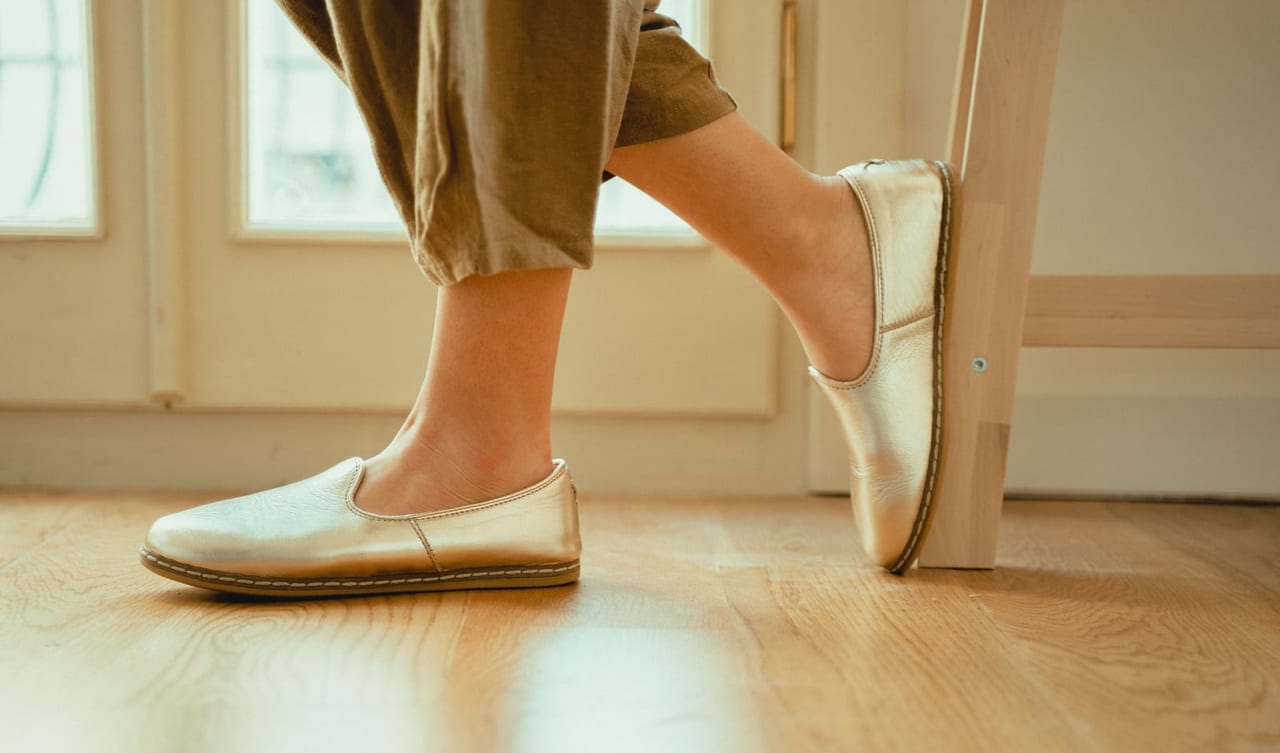Feet in gold leather slip-on shoes on a light wooden floor, one heel raised