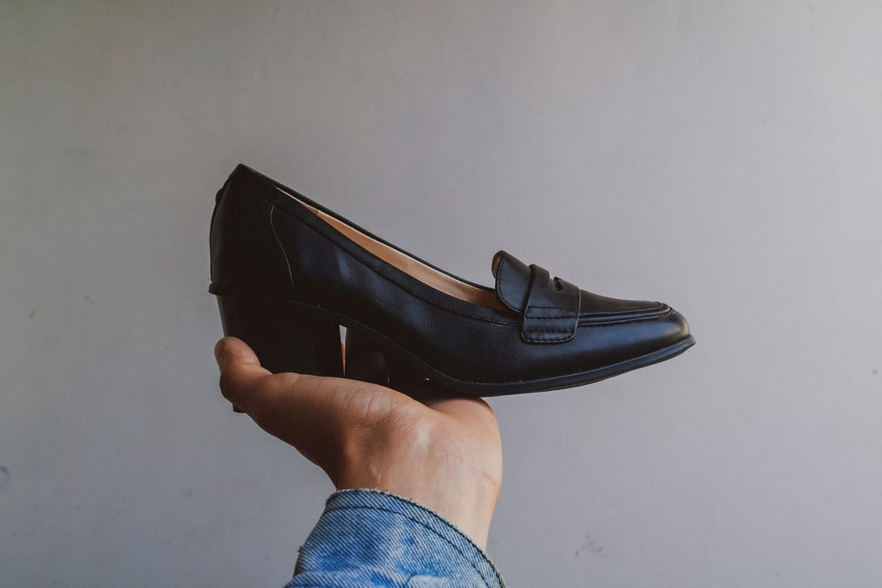 Hand holding up a black leather heeled loafer against a plain gray wall