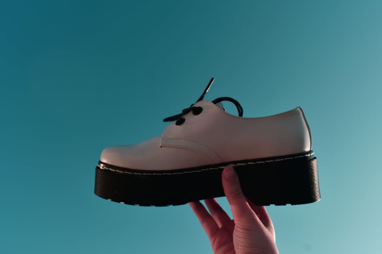 Hand holding a pale lace-up shoe with a thick black sole against a teal background