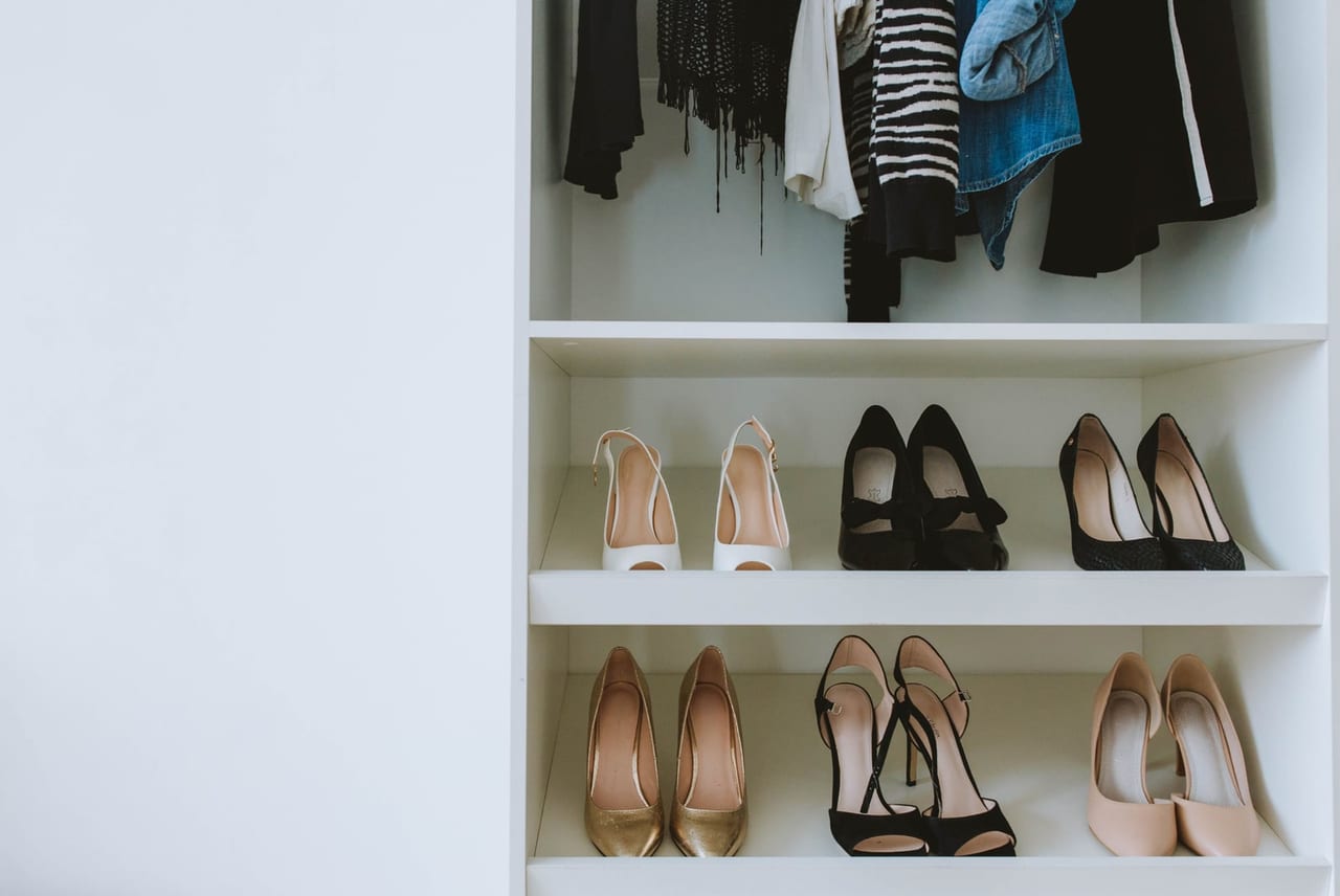 Six pairs of heels lined up on white closet shelves below hanging clothes