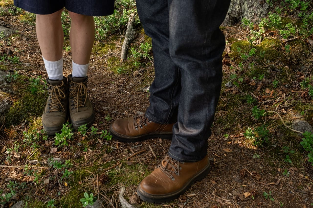 Two people in leather hiking boots standing on a mossy forest floor, one in white socks