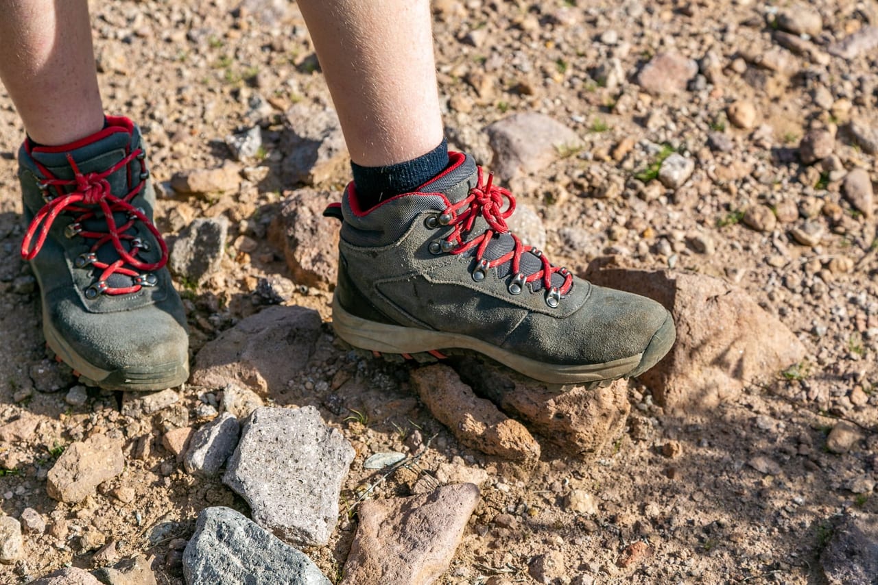 Gray suede hiking boots with red laces standing on dry, rocky dirt in sunlight