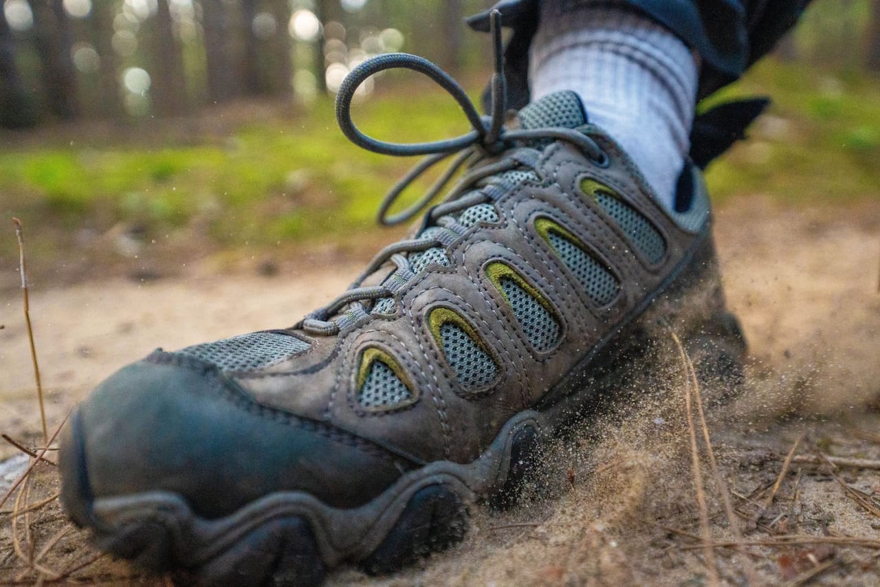 Gray hiking shoe and white sock kicking up sand mid-step on a forest trail