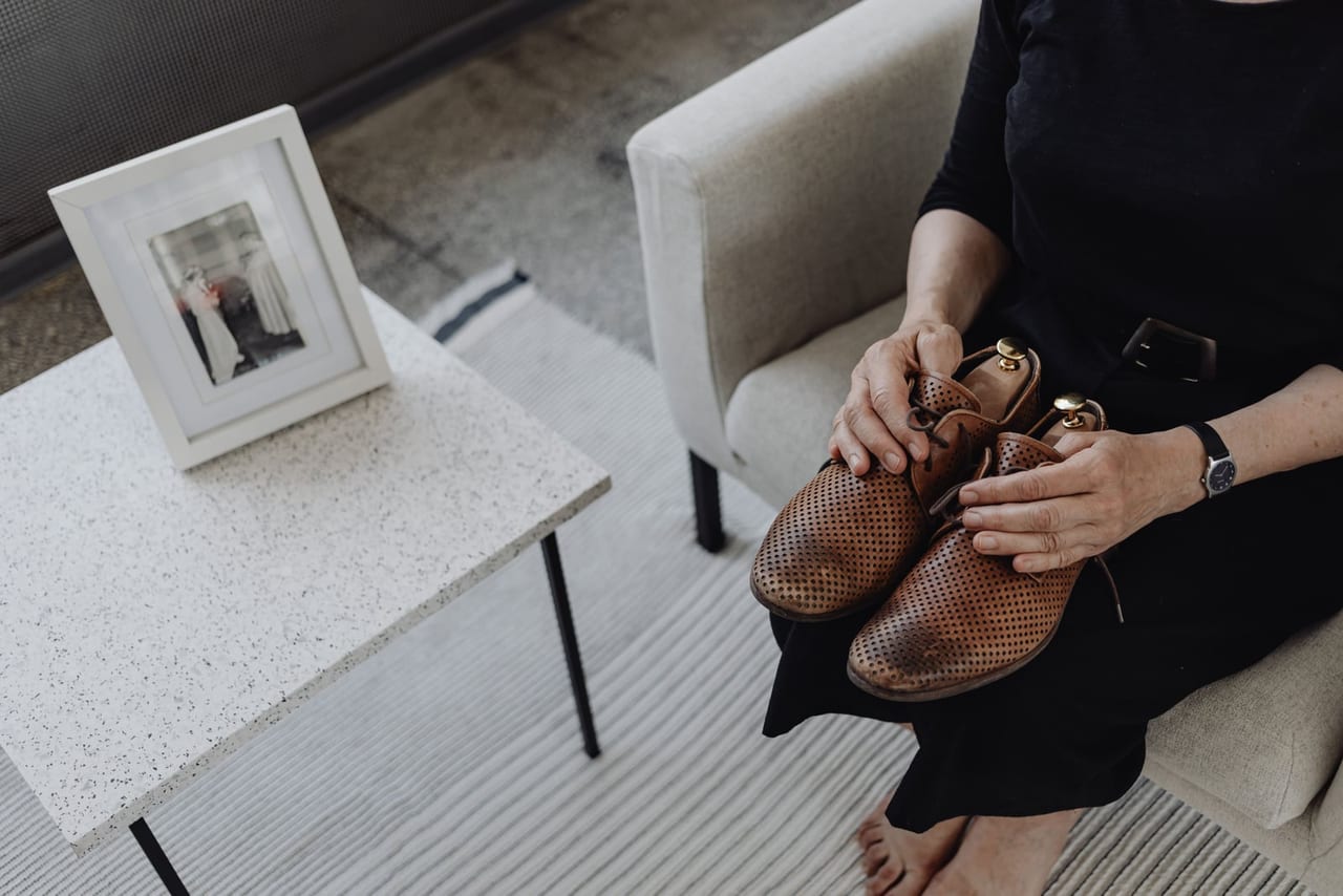 Barefoot woman in black sitting in an armchair holding a pair of woven brown leather shoes