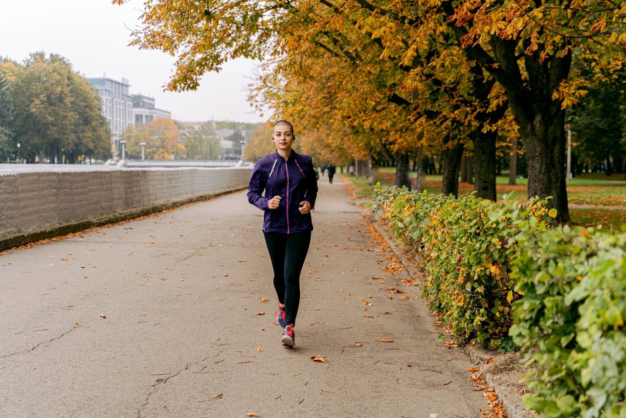 Woman in a purple jacket jogging along a paved, tree-lined path scattered with autumn leaves