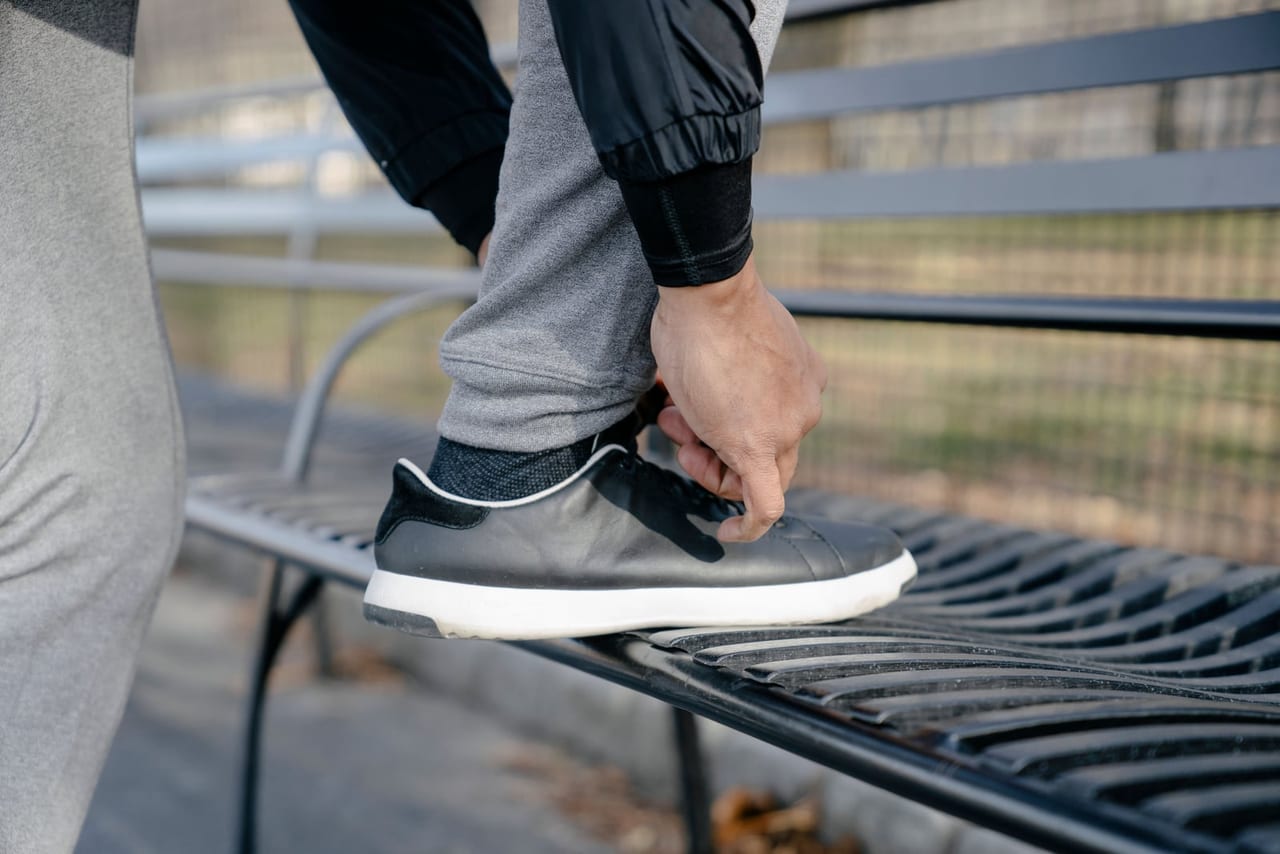 Hand adjusting the laces of a black leather sneaker with a white sole on a bench