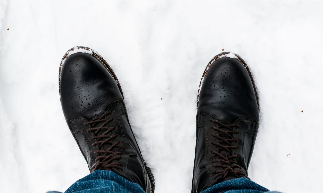 Looking down at black leather lace-up boots beaded with water, standing in fresh snow