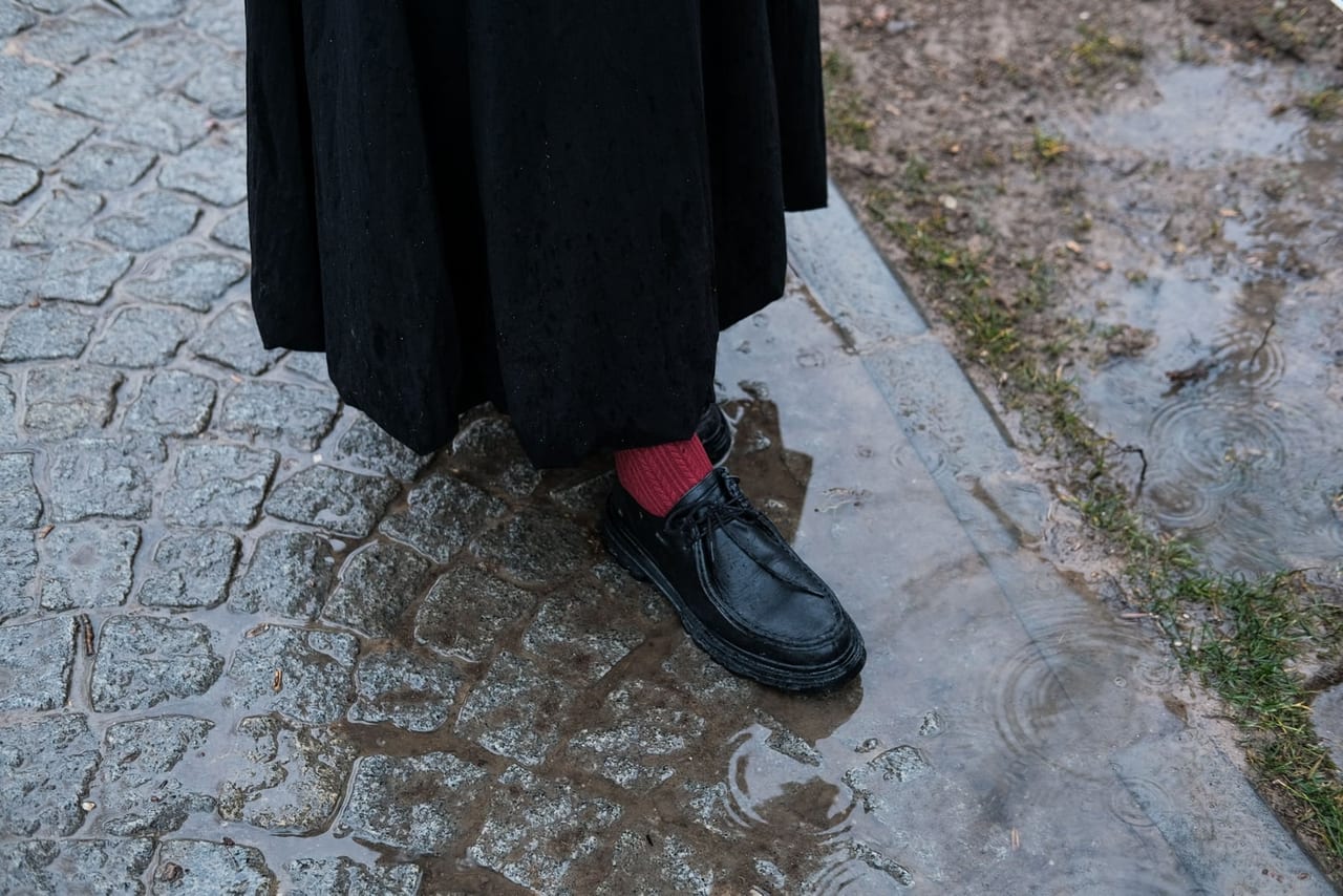 Black leather shoe and red sock on wet cobblestones beside a rippling puddle