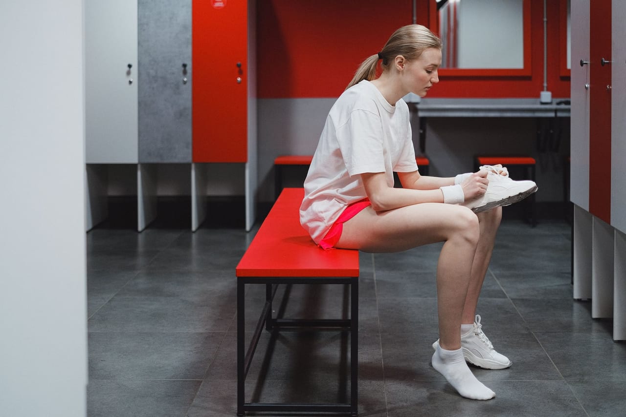 Woman on a red locker room bench putting on white sneakers over ankle socks